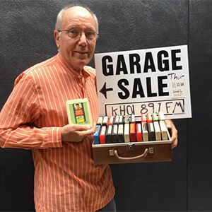 Bob holds a case of 8-track tapes and a garage sale sign with event details and a radio station name.