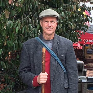 Ed Fallon stands outdoors holding a walking stick, with greenery and a red bookstore sign in the background.