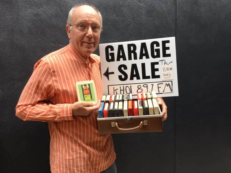 Bob holds a case of 8-track tapes and a garage sale sign with event details and a radio station name.