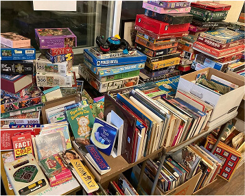 A table crowded with stacks of board games, jigsaw puzzles, and books.