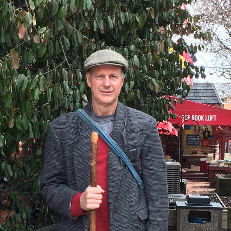 Ed Fallon stands outdoors holding a walking stick, with greenery and a red bookstore sign in the background.